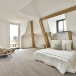 Interior of spacious light contemporary bedroom furnished with comfortable bed placed on parquet floor near white sloped uneven wall with decorative beams and window